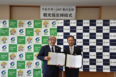 さぬき市とJAF香川支部の観光協定締結式で担当者2人が協定書を手に並んでいる写真