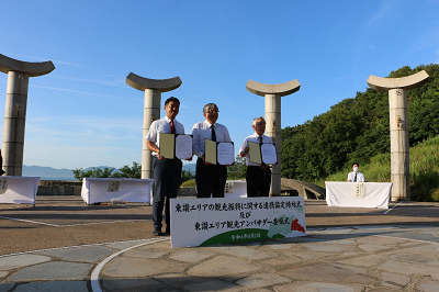 石碑が並ぶ広場でスーツ姿の男性3人が認定書を掲げて記念撮影している写真