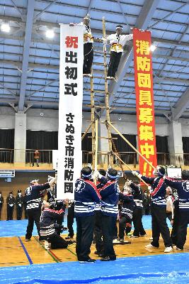 複数人で支えるはしごに2人で乗り、頂上付近から団員募集と出初式を祝う垂れ幕を掲げる様子