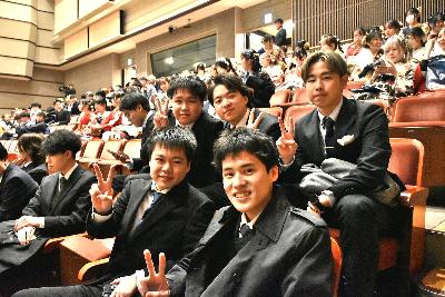 会場内の席に座りながらピースをする5名のスーツ姿の男性