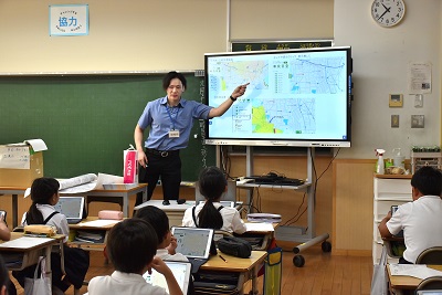 電子黒板に指さす市職員とタブレット端末を前に説明を聞く児童