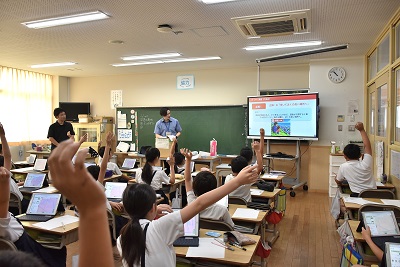 防災クイズを出す市職員と手を挙げて答える児童
