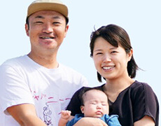 赤ちゃんを抱き、青空のもとで幸せそうに微笑む移住者夫婦