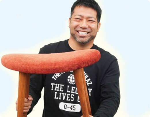 赤い座面の椅子を持ち、満面の笑みを浮かべる移住者の男性