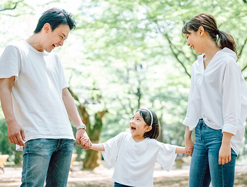 手をつなぎ、笑顔で見つめ合う親子三人と新緑の背景
