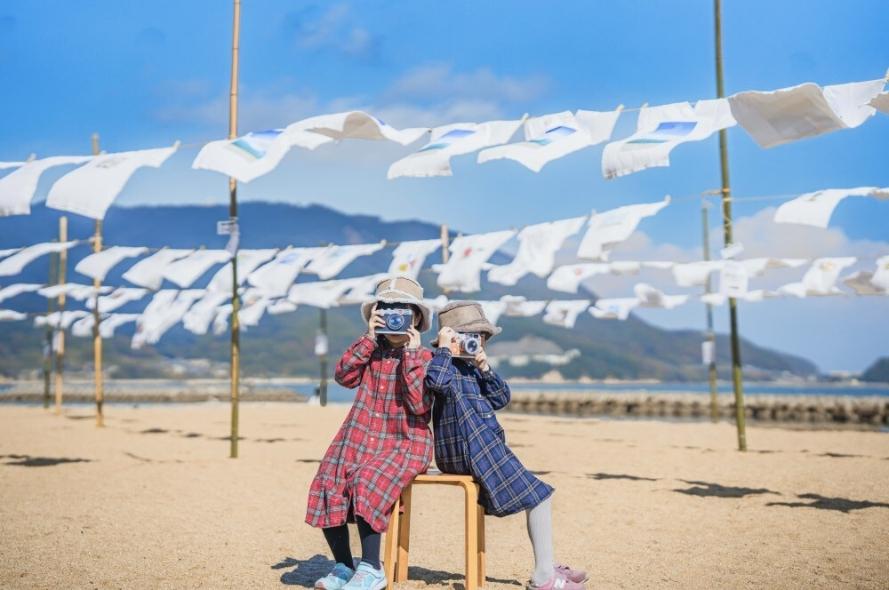 風に揺れる白いTシャツを背景に、砂浜でカメラを構える二人の子ども
