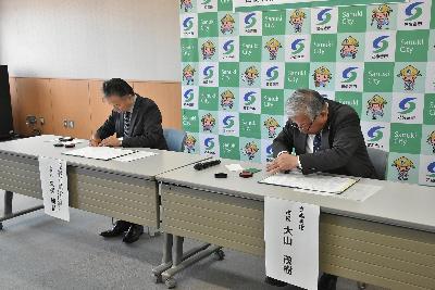 大山市長と久保取締役が調印書に署名する様子