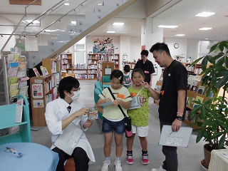 白衣を着たスタッフから出題された謎解きゲームに親子探偵がチャレンジしている様子の写真