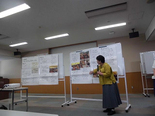 黄色の上着を着た女性がホワイトボードに資料を掲示して男女共同参画社会につながる活動の実践報告をしている写真