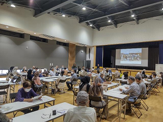 公民館で多くの参加者が机と椅子に座りスクリーンの映像を見ている写真