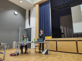 壇の前で女性がマイクを持って話しもう一人の女性がテーブルについている写真