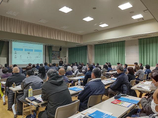 多くの参加者が会場の椅子に座りスクリーンを見ている写真