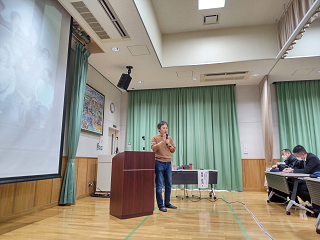 講師が大きなプロジェクターの前で演壇に立ち受講者に向けて講演している写真