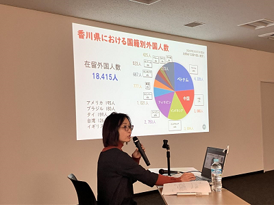 スクリーンに「香川県における国籍別外国人数」と書かれたスライドが表示され、それについて説明している講師の写真