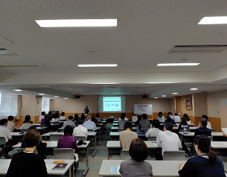 多くの参加者が奥のスクリーンや手元の資料を見ながら講演を聞いている写真