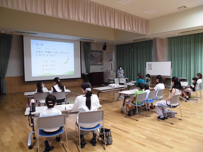 スクリーンに映し出された内容の説明を多くの子どもや参加者が聞いている写真