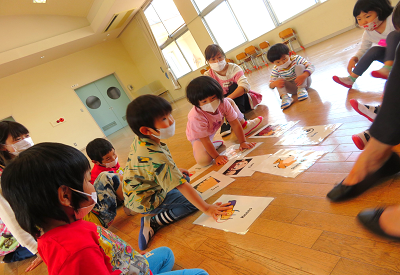 ホールで子どもたちが床に並べたカードを囲んで英語のカード探しゲームをしている様子の写真
