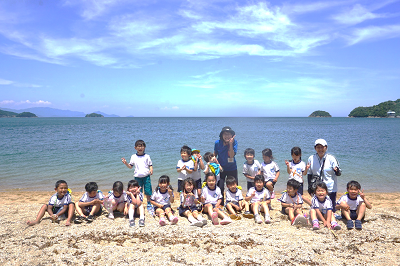 海辺の砂浜で園児たちと先生が並んで座り青い海を背景に記念撮影している様子の写真