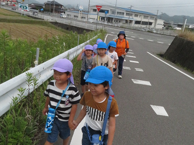 薄紫色や水色の帽子を被った園児がガードレール沿いを2列で歩いている写真