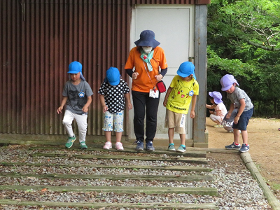 薄紫色や水色の帽子を被った園児たちが先生と屋外で遊んでいる様子の写真