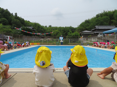 プールサイドに座り、黄色い帽子をかぶった子どもたちが先生の話に耳を傾けている様子の写真