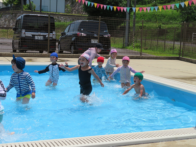 プールで子どもたちが先生と一緒に水に入り、追いかけっこをして楽しく遊んでいる様子の写真