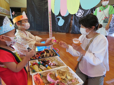 屋内で保護者に紙で作られた食べ物を渡している園児たちの写真