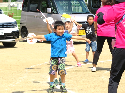 屋外でうちわを持って両手を広げながら踊る複数の園児の写真