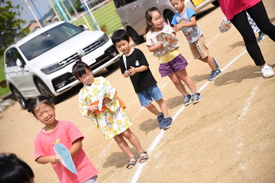 屋外で浴衣やシャツを着た複数の園児がうちわを持って並んで踊っている写真