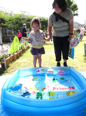 保護者と手をつなぎ方手に黄色い網を持ち目の前にあるヨーヨーの浮いたビニールプールをみつめる園児の写真