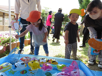 帽子を被った園児たちがビニールプールに浮かぶヨーヨーを網で掬おうとしている様子の写真