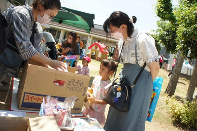 屋外で段ボールから商品を取り出す女性とその様子を保護者と見守る園児の写真