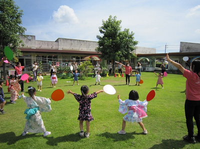 芝生の上でうちわを持って両手を広げて輪になり踊る園児たちの写真