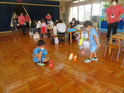 教室の中で椅子に座り工作をする児童たちと出来上がった作品で遊ぶ園児たちの写真