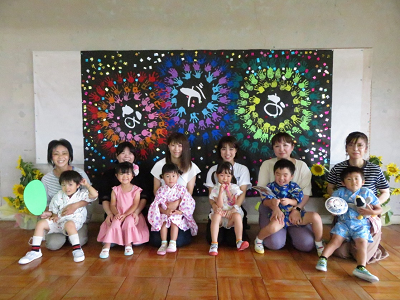 子どもたちの手形で作った大きな花火のアート作品を背景に記念撮影をする園児と保護者たちの写真