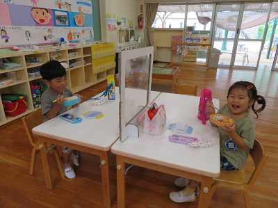 教室でパーティションに区切られた向かい合った机で椅子に座り、笑顔でお昼ご飯を食べる男の子と女の子の写真