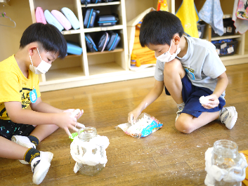 教室に座って、集中した様子でビンに思い思いに紙粘土を付ける2人の男の子の写真