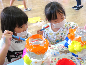 二人の女の子が紙粘土を貼ったビンに鮮やかなオレンジや黄色、ピンクなどの色を筆で塗っている写真