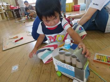 牛乳パックにペットボトルの蓋でタイヤを付けた手作りの車に、屋根を付けている男の子の写真