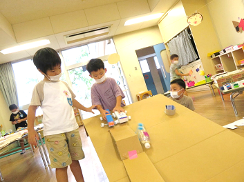 教室に段ボールで作られた坂道で手作りの車を走らせて遊んでいる3人の男の子の写真