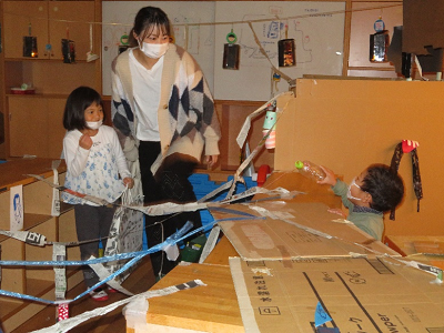 室内の段ボールの基地のそばで保護者と子供が遊びの様子を見守っている写真
