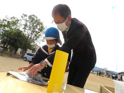 園庭のテーブルで園児が先生に見守られながら焼き芋をアルミホイルで包んでいる写真