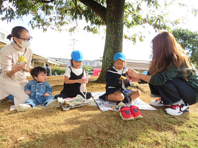 木陰の芝生で保護者と園児たちが座って焼き芋を食べている写真