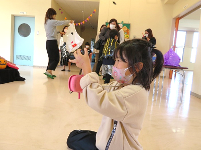 室内でマスクをした女の子が膝をついてカメラで撮影している写真