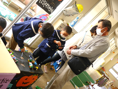 室内で園児が作品展示のテーブルを覗き込んでいるのを保護者が見守っている写真