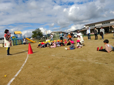 園庭で先生の話を聞きながら園児たちが地面に座って集まっている写真
