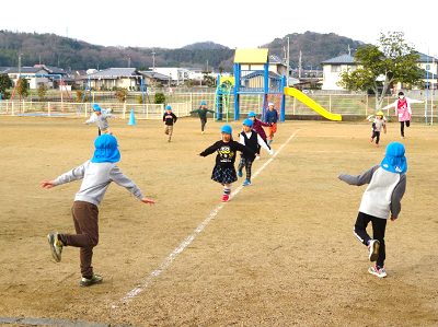 屋外で青い帽子の園児たちが体を動かして遊んでいる写真