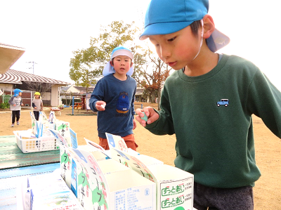園庭で青い帽子の園児がテーブルに並べられた牛乳パックの容器にペットボトルの蓋を入れている写真