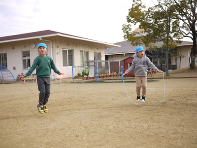 園庭で青い帽子の園児二人が縄跳びを同時に跳んでいる写真