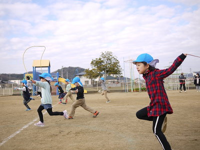 園庭で青い帽子の園児たちが列になって縄跳びをしながら走っている写真